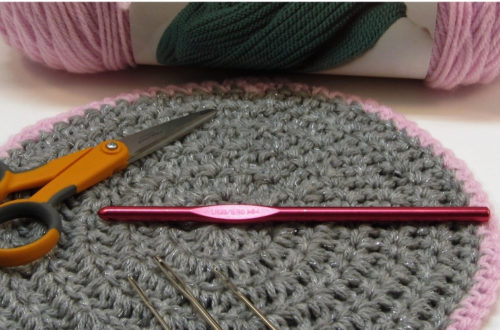 Crochet hook, darning needles, small scissors and yarn on a round crocheted doily,.