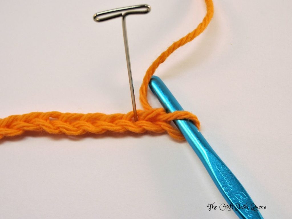 Orange yarn crocheted in a chain.  Blue crochet hook.  T-pin indicating the second chain from crochet hook.